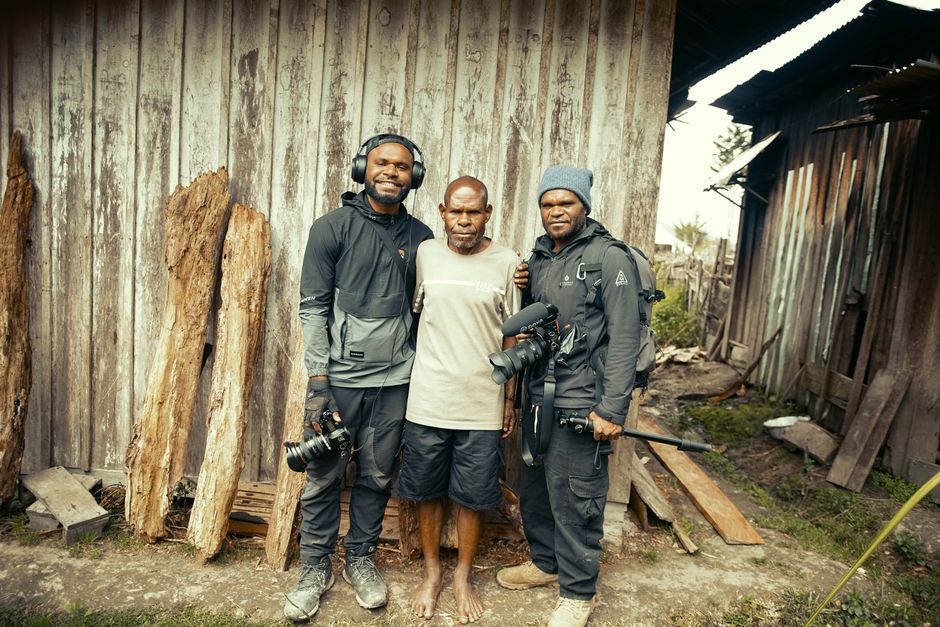 Turismo Sustentável — Three men, two with cameras, pose in front of a rustic wooden building outdoors.